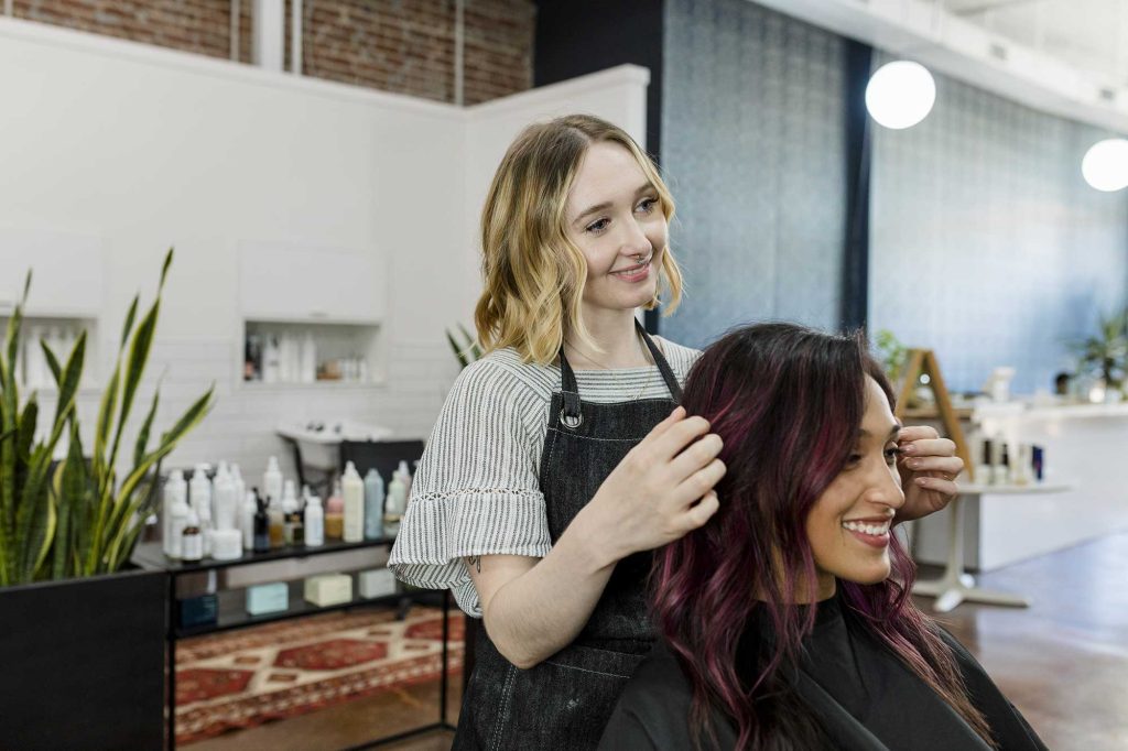 A hairstylist with blonde hair smiles while cutting a woman's dark hair with purple highlights in a modern salon. The mood is friendly and relaxed.