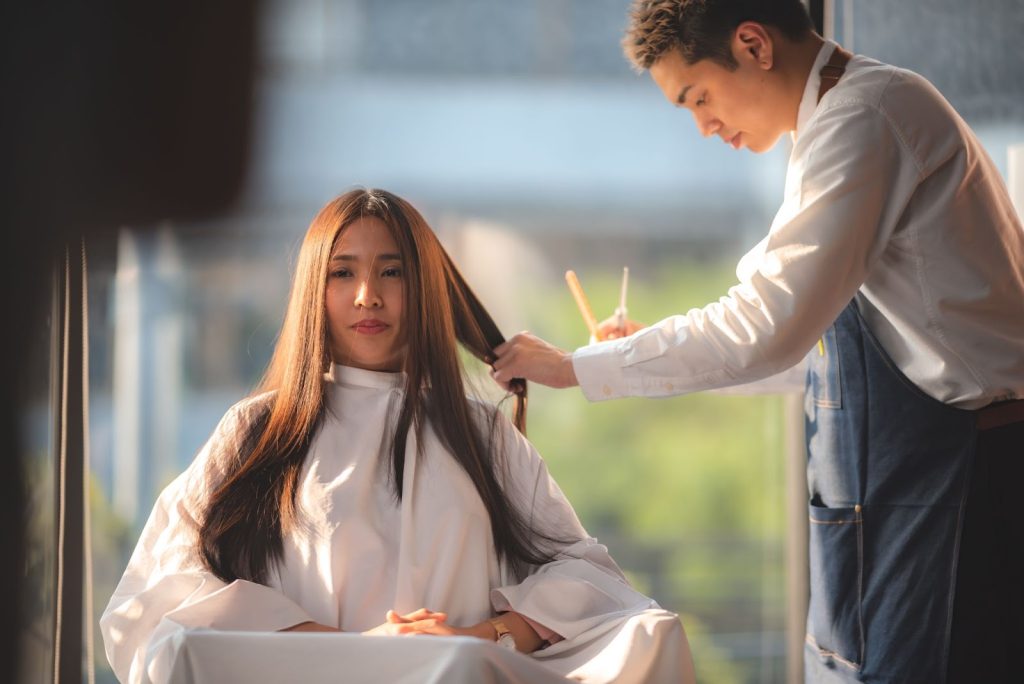 A woman with long hair sits in a salon chair, wearing a protective cape. A hairstylist in a white shirt and apron carefully trims her hair in a well-lit room.
