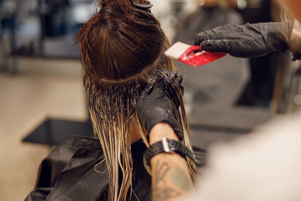 Hairdresser wearing black gloves applying hair dye to long brown hair in a salon.