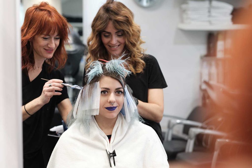 Two hairstylists apply hair color to a seated client's hair, creating a vibrant blue look. The client wears a white cape and has a subtle smile. The salon is modern with shelves in the background.